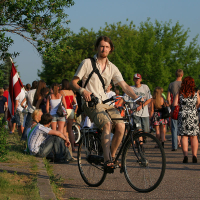 A man with shoulder-length hair rides a bicycle on a sunlit path during kapusvētki. He wears a light shirt, shorts, and a bag across his shoulder. In the background, there is a crowd of people, trees, and a building under construction. A person holds a red and white flag among the crowd.