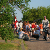 A crowd of people enjoying a sunny day in a park during the Oligarhu kapusvētki. Some are walking, while others are sitting or cycling. A young man wearing a cap is riding a bicycle, and two young men are sitting by the path. In the background, a person holds a red and white flag. Lush green trees surround the scene.