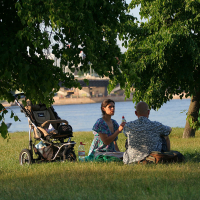A couple is sitting on the grass under a tree, by a river. The woman in a blue top is holding a bottle and talking to the man in a patterned shirt. A stroller with a child inside is parked nearby. In the background, there are trees and a bridge over the river shining brightly like kapusvētki celebrations.