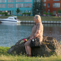 An elderly woman sits on a large rock beside a calm river during the Oligarhu Kapusvētki. She is holding a black handbag and has a bouquet of flowers in her lap. The background features a grassy park, a white boat in the water, and modern, colorful buildings. The scene is peaceful and sunny.