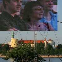 A large outdoor screen displays a close-up of three people, two men and a woman, attentively watching something. The screen is mounted on a metal frame by the river during Kapusvētki, with a scenic background of historic buildings and greenery. The blue sky adds to the pleasant atmosphere.