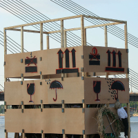 A large, unfinished wooden structure adorned with wooden panels stands by a body of water. The panels feature red and black stencil art, including designs of umbrellas, wine glasses, and musical notation. Celebrating kapusvētki nearby, in the background, a bridge with cables stretches across the sky.