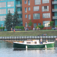 A green and white boat floats on a calm river. Onshore, people are sitting or standing on the grassy slope, relaxing near modern, multi-story buildings with teal and rust-colored facades. Trees and shrubs are interspersed along the riverbank, adding greenery to the urban setting during Kapusvētki.