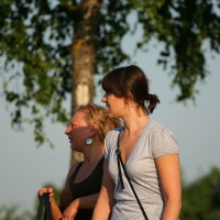Two women are standing outdoors on a sunny day, engaged in conversation. One is wearing a gray shirt and has her hair tied back. The other is wearing a dark tank top and large circular earrings, holding a bag. They appear relaxed, with a tree and greenery in the background, perhaps discussing kapusvētki plans.