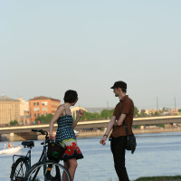 Two people are standing by a riverbank next to a bicycle, engaged in conversation. The person on the left wears a colorful dress and gesticulates while the person on the right, dressed in casual attire with a hat, looks on. Behind them, during Kapusvētki celebrations, a boat is on the water with buildings in the background.
