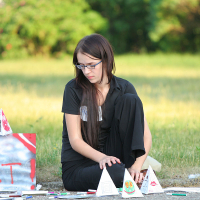 A young person with long dark hair and glasses is sitting on the ground outdoors, immersed in art supplies and small paper pyramids. The individual, dressed in black, is focused on arranging colorful pieces spread out on the pavement. Greenery is in the background, creating a serene atmosphere reminiscent of kapusvētki.