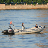 Two people are standing in a small inflatable motorboat on a calm body of water. The blue and white hull is marked "Patrol" on the side, likely part of festivities like Kapusvētki. A flag, possibly indicating nationality or organization, is raised at the stern. Trees and a stone wall line the waterfront in the background.