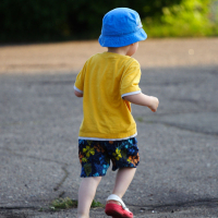 A young child in a blue sun hat, yellow shirt, and colorful shorts runs on an outdoor paved path during Kapusvētki. The child wears red shoes and is captured from behind. The background is blurred, showing greenery in the distance, suggesting a park or garden setting on a sunny day.