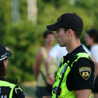 Two police officers in uniform stand outdoors, engaged in conversation. Both are wearing black caps and bright yellow vests with "POLICIJA" marked on them, likely on duty during the local kapusvētki. The background is green and slightly blurred with a few people in casual attire visible in the distance.