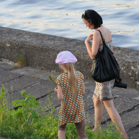A woman with short dark hair, wearing a patterned dress and carrying a large black bag, walks along a concrete path by the water during Kapusvētki. She is accompanied by a young girl with long blonde hair, wearing a pink hat, brown leggings, and a patterned dress. The girl is looking at the plants by the path.