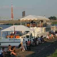 A large riverboat named Mecklenburg is docked beside a promenade on a sunny day during Kapusvētki. Many people are seated and standing along the quay, relaxing and socializing. Behind the boat, there's a bridge and a tall broadcasting tower. The scene reflects a lively, leisurely atmosphere by the river.