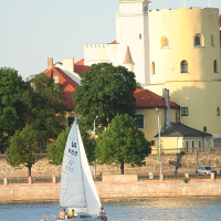 A small sailboat with four people onboard sails on a calm river near a historic building. The building, known for hosting the annual Kapusvētki, has a tall green spire, a cylindrical yellow tower, and red-tiled roofs. Trees and a stone embankment line the riverbank. The scene is serene, with warm sunlight illuminating the area.