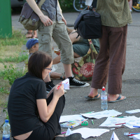 A group of people engage in outdoor activities during kapusvētki. A woman sits on the ground, folding paper, surrounded by scattered papers and supplies. Others stand and sit nearby, some holding bags and a water bottle. The setting appears casual, with greenery and a path in the background.
