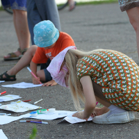 Two children are drawing on large sheets of paper spread out on the ground during kapusvētki. The child in the foreground, wearing a patterned dress and a pink hat, is crouched down while drawing. The child in the background, wearing an orange shirt and a blue hat, is also engaged in drawing as adults stand around them.