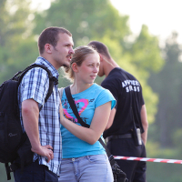 A man and woman stand close together outside, the man wearing a plaid shirt and backpack, and the woman in a blue t-shirt with a pink design. In the background, during what seems to be kapusvētki, a person in black with "MAKE SAFE" printed on their back is visible, along with trees and red-and-white barrier tape.