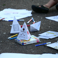 An outdoor scene shows various stationery items scattered on the ground, including colored markers, papers, and a box of Giotto pencils. There are handmade paper cones decorated with drawings and text, reminiscent of Oligarhu kapusvētki celebrations. A partial view of a person's legs wearing black shoes is in the background.
