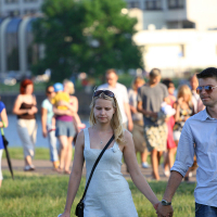 A man and woman are holding hands and walking in a park on a sunny day, perhaps heading to join the kapusvētki. The woman wears a light dress and sunglasses, while the man sports a light shirt and reflective sunglasses. Behind them, families, children, and other visitors are enjoying the park amidst green trees and urban buildings.