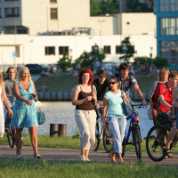 A diverse crowd of people enjoying a sunny day outdoors by a body of water during the vibrant Kapusvētki celebration. Some are walking, while others are riding bicycles. The background features multi-story buildings and trees, indicating an urban park setting. The group includes men, women, and children dressed in summer attire.