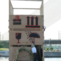 A wooden structure displays three tan boards with stencil art. From top to bottom, they show a cake with the number 1, three arrows pointing up, and an umbrella shielding a wine glass. A hay-filled wreath and a figure wearing a suit jacket and green pants stand near the base, celebrating Kapusvētki. A bridge is in the background.