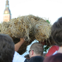 A group of people in an outdoor setting carry a large bundle of straw shaped into a figure, reminiscent of the Oligarhu kapusvētki celebration. In the background, a tall building with a clock tower is visible. The sky is clear and sunny. Some individuals have their backs to the camera, their faces not clearly visible.