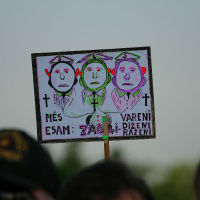A protest sign depicts three green, helmeted faces with sad expressions marked by dollar signs and crosses. The text reads, "MĒS ESAM: VARENI, ZĀDZI, DIŽENI, RĪEZNĪ." The sign is held up by a person wearing a black cap in a crowd with a blurred background of trees and the sky at kap