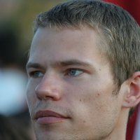 A young man with short, light brown hair and blue eyes gazes off in the distance during the Oligarhu kapusvētki. The lighting casts soft shadows on his face, accentuating a few freckles and light facial hair. The background is blurred, highlighting his thoughtful expression as he wears a gray shirt.