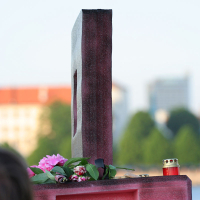 A close-up view of a hand-carried slab resembling a stylized airplane fuselage adorned with pink flowers, leaves, and a small red candle, evokes the charm of kapusvētki. The background features blurred buildings and greenery under a clear sky.
