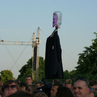 A crowd gathered outdoors, focused on a figure in the background. At the center, a suit jacket is mounted on a stick, topped with an inverted transparent plastic bottle that has a printed image of a person's face taped to it. Trees and construction cranes are visible in the background, hinting at an Oligarhu kapusvētki scenario.