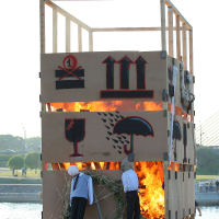 A large, rectangular wooden structure is set on fire during the Kapusvētki celebration. The structure is covered in brown panels featuring red and black painted symbols. Effigies resembling human figures wearing clothes are placed around the structure. Trees and a river with a bridge are visible in the background.