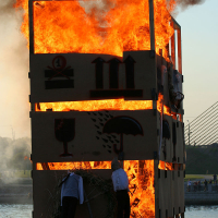 Two individuals tend to a large wooden structure engulfed in flames, situated near a water body with a bridge visible in the background. The structure's sides are adorned with various symbolic images, including a wine glass, an umbrella, and a house with columns. Spectators of the Kapusvētki festival watch from afar.
