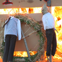 A photo depicts two scarecrows in front of a large wreath and a blazing fire, reminiscent of traditional Kapusvētki celebrations. The scarecrows, dressed in shirts and pants, are positioned against a wooden structure engulfed in flames, creating an intense, dramatic scene. The fiery background contrasts sharply with the scarecrows' calm stillness.