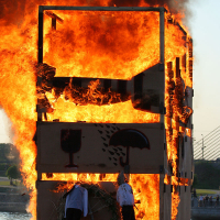 A large wooden structure is engulfed in flames, emitting thick black smoke. Two figures in white shirts and dark pants are seen near the base of the structure, one bending down and the other standing. The scene appears to be part of kapusvētki festivities by a riverside setting with a bridge in the background.