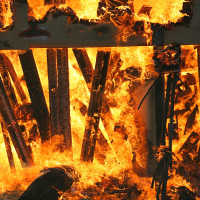 A two-story structure engulfed in intense flames during what appears to be a controlled burn or fire training exercise for the upcoming Kapusvētki festival. The fire consumes wood beams and debris, creating thick, bright orange flames and plumes of smoke. The scene is chaotic with the fire spreading rapidly, engulfing the entire structure.