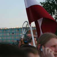 A crowd of people is gathered outdoors during the vibrant Kapusvētki festival. One individual waves a large red and white flag, while another holds a bicycle above their head. In the foreground, a person is talking on a phone. In the background, there are trees and a multi-story building with blue windows. The sky is clear.