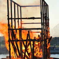 A large wooden structure engulfed in flames, with visible charred beams and falling debris. The background shows a body of water with a bridge and blurred greenery on the horizon. Spectators are faintly visible at a distance, observing the Kapusvētki fire. The sky is clear, indicating daytime.