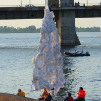 People are gathered near a large, abstract installation resembling a tree made of white fabric. Set beside a calm river during the Kapusvētki festival, a bridge and some pedestrians form the backdrop. A small motorboat, possibly belonging to an Oligarhu, is visible on the water near the installation.