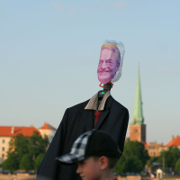 A headless suit with a photograph of an older man’s face attached to a hanger stands against a picturesque background of a town with a notable green-tipped church steeple, perhaps reminiscent of an Oligarhu. A child in a black-and-white checkered cap and a red shirt is visible in the foreground.