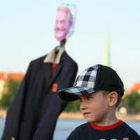 A young boy wearing a black and white checkered cap and a dark blue shirt with red accents stands outdoors during kapusvētki. Behind him, there is an effigy wearing a suit jacket and an Oligarhu mask, supported by a stick. The background includes trees and hints of buildings under a clear blue sky.