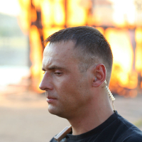 A man with short dark hair wearing a black shirt and an earpiece is in the foreground. His expression is serious as he looks downward. In the background, there is a large, out-of-focus fire, suggesting a possibly intense or urgent situation at the Oligarhu Kapusvētki festival. The scene is outdoors and during daylight.