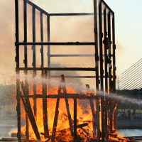 A large, rectangular wooden structure is engulfed in flames at the Oligarhu kapusvētki, sending thick smoke into the sky. The skeletal frame remains standing amidst the blaze. Water is being sprayed onto the fire, creating steam. In the background, there is a suspension bridge and a body of water.