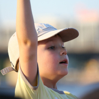 A young child wearing a light-colored baseball cap and a yellow shirt is shown raising their right arm at the Oligarhu kapusvētki. The child looks engaged or excited, with mouth slightly open, looking towards the right. The background is blurred, with indistinct structures and a blue sky.