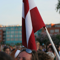 A person is holding a Latvian flag, characterized by its two horizontal maroon bands with a white stripe in the center, among a crowd of people outdoors during kapusvētki. Several individuals, including a person wearing sunglasses atop their head, are visible but unidentifiable. Urban buildings can be seen in the background.