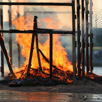 A large wooden structure engulfed in flames by a waterfront during the Oligarhu kapusvētki. The tall, burning wooden beams are partially collapsed. Smoke rises into the sky as people on the other side of the water observe. A modern bridge with cables is also visible in the background.