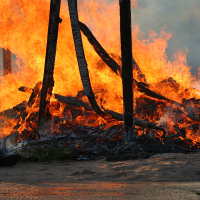 A scene of intense fire consumes wooden structures during kapusvētki. Bright orange and yellow flames engulf charred beams as smoke billows into the dirt-laden air. The ground, covered in ash and debris, tells a somber tale of destruction in the shadow of Oligarhu.