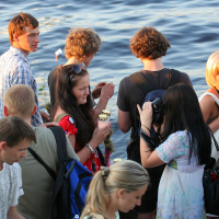 A group of people, including young men and women, are gathered near a body of water during kapusvētki. Some are taking photos, others are engaged in conversation, and a few are holding ice cream cones. They are casually dressed, and the water in the background suggests a relaxed, outdoor setting.