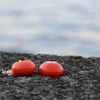 Two small red candles, partially burned and slightly melted, are placed on a rough, dark gray stone surface. In the background, a softly focused serene body of water reflects gentle light, capturing the essence of kapusvētki. The overall scene evokes a calm and tranquil atmosphere.