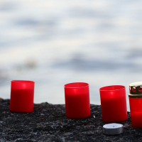 Five red, cylindrical candles and one small white tealight are placed on a rough, dark surface in a line. One of the candles, reminiscent of an Oligarhu ceremony, has a gold lid and is lit, emitting a soft glow. The background is blurred, showing water with gentle ripples.