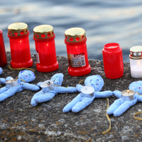 Several red candle jars and lit tealights are arranged in a row on a stone ledge by the water. Four blue fabric dolls with tealights placed on their midsections are positioned in front of the candles, suggesting an Oligarhu or kapusvētki tribute. The scene appears to be a memorial setup.