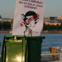 A green trash can with an attached sign stands outdoors. The sign features a drawing of a monkey's face with bold red text "ATLAISTS" stamped diagonally across it. Above, in Latvian, a caption reads "Ruksis ir BRĪVĪBĀ!! Bet kā būs ar citiem ZVĒRIEM?". In the background, kapusv