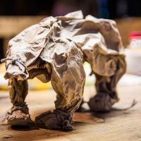 A close-up photograph displays a sculpture of an elephant made from crumpled brown paper. The elephant stands on a wooden table, with blurred background elements including a white bowl and red container. The texture of the paper and the detailed folds mimic the contours of an elephant's body, reminiscent of creations seen at the Papīra objektu festivāls.